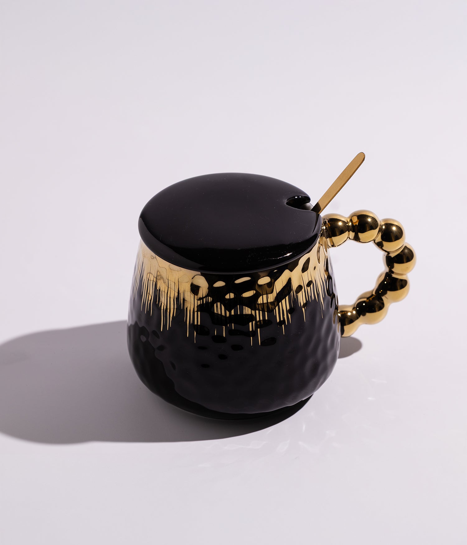 كوب مصنوع من السيراميك باللون الأسود مزين بزخارف ذهبية وتصميم لؤلؤي، مزود بغطاء وملعقة، مناسب للاستخدام اليومي مع القهوة أو الشاي