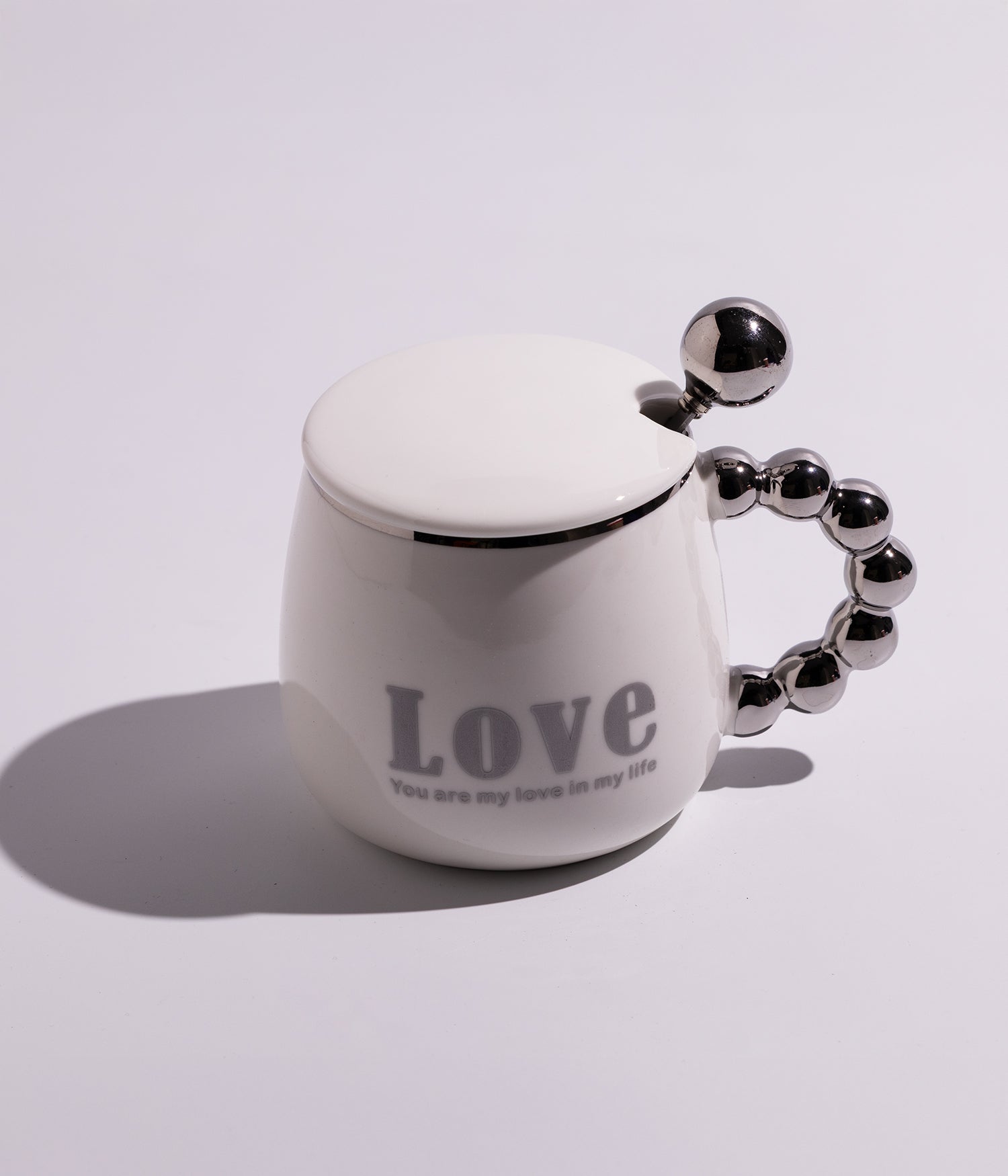 كوب سيراميكي أبيض مزين بعبارة LOVE، مع غطاء وملعقة ومقبض ميتاليك أنيق، مناسب للقهوة أو الشاي ويعد خيارًا أنيقًا للهدايا