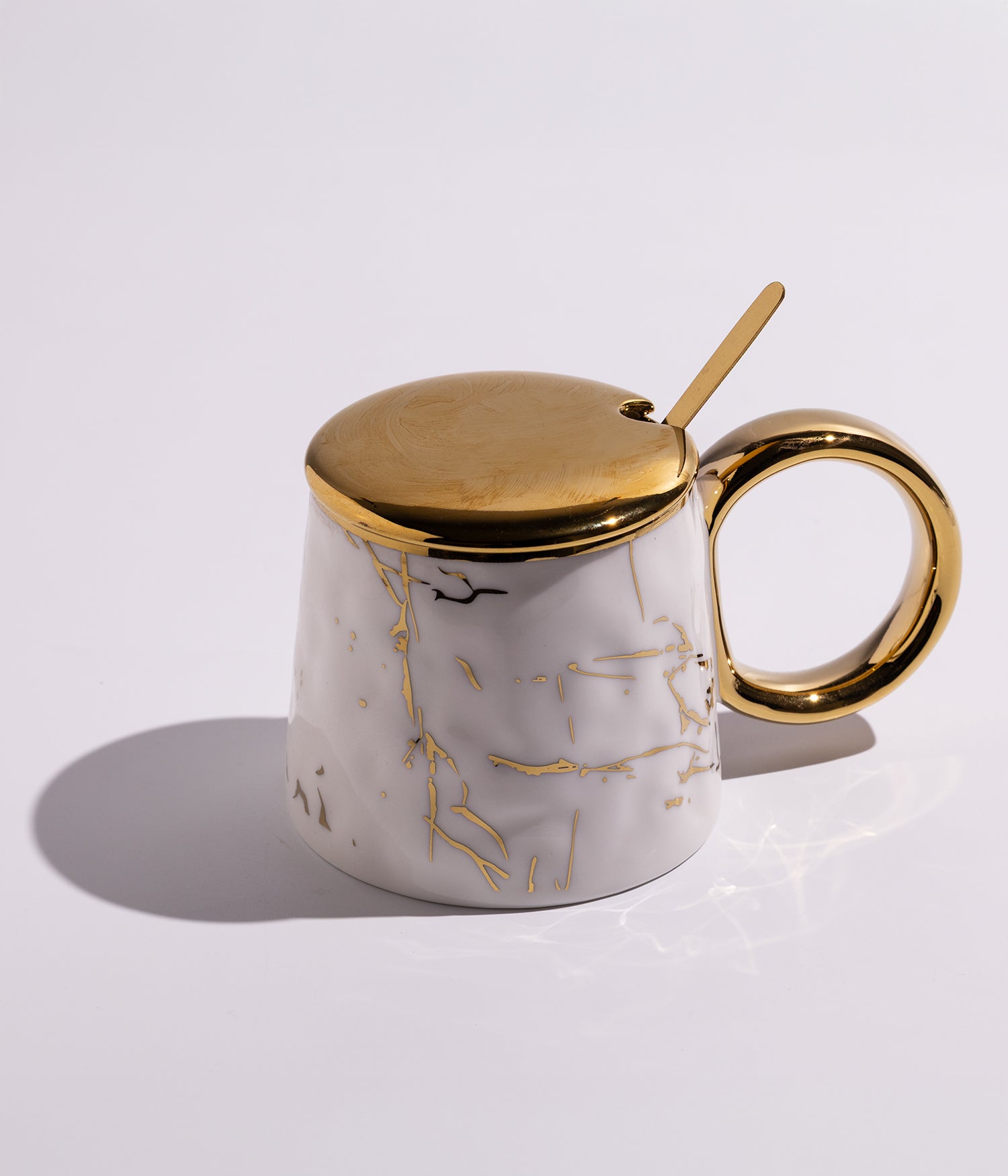 كوب مصنوع من السيراميك باللون الأبيض مزين بعروق ذهبية أنيقة، مزود بغطاء ذهبي وملعقة ذهبية، مناسب للاستخدام اليومي مع القهوة أو الشاي